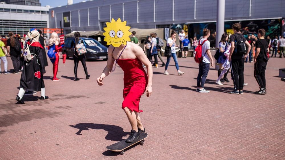 Photo of a person in red dress, on a skateboard. The person's face is covered by a drawing of a smiling sun. Walking or standing people and a fragment of a building as a background.