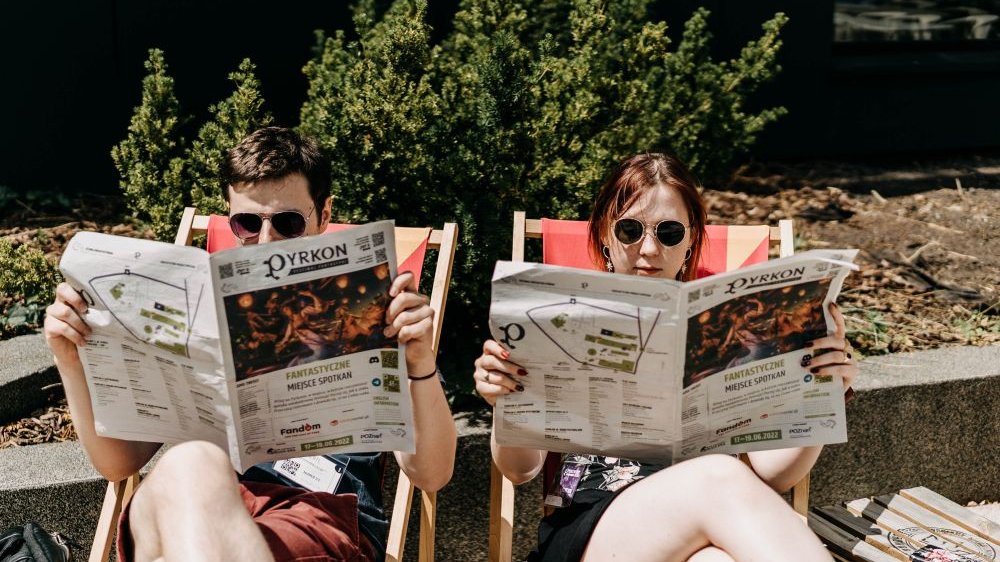 A man and a woman sitting on deck chairs and reading Pyrkon newspapers.