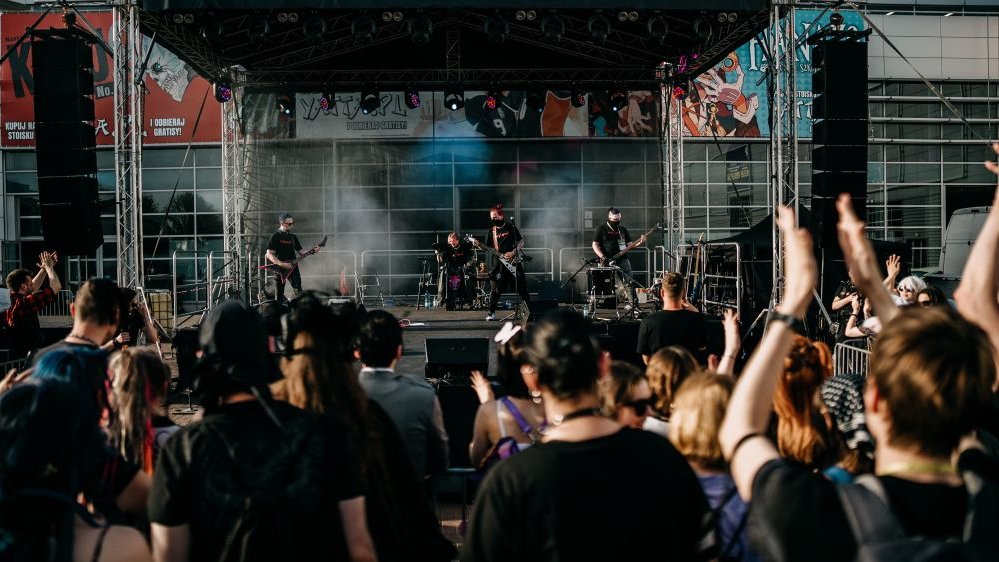 Photo of a music band (three guitarists and one drummer) performing on stage. People watching the concert in front of the stage.