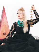 Photograph shows the artist. The woman is wearing a black dress. The sleeves are made of lace and the bottom is made of tulle. Light straight hair falls to the left shoulder. The artist raises her head slightly and looks beyond the frame. She is accompanied by musical instruments. White background.