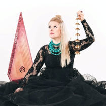 Photograph shows the artist. The woman is wearing a black dress. The sleeves are made of lace and the bottom is made of tulle. Light straight hair falls to the left shoulder. The artist raises her head slightly and looks beyond the frame. She is accompanied by musical instruments. White background.