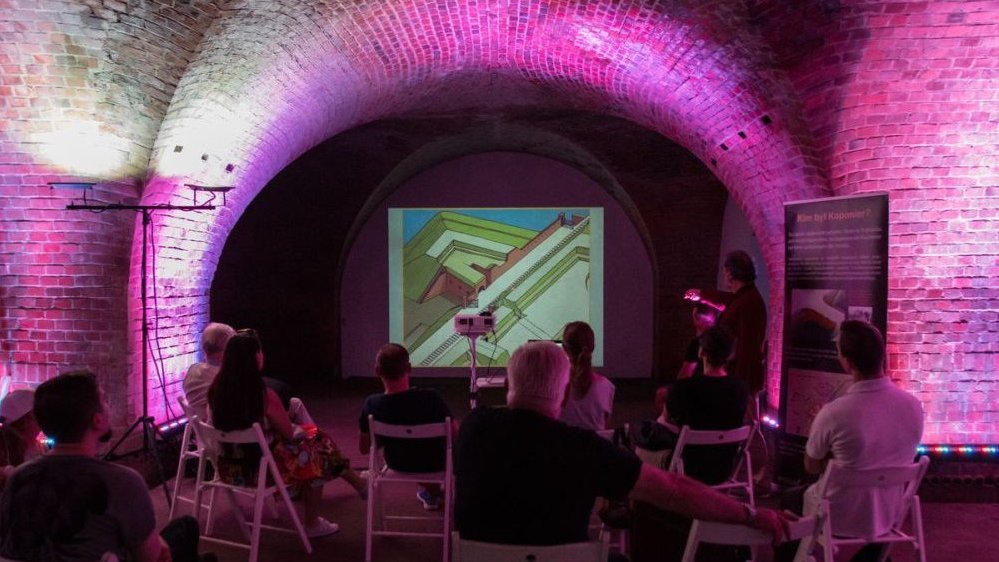 Photo of an interior of a fort - a few people sitting on chairs and watching a presentation on a screen.