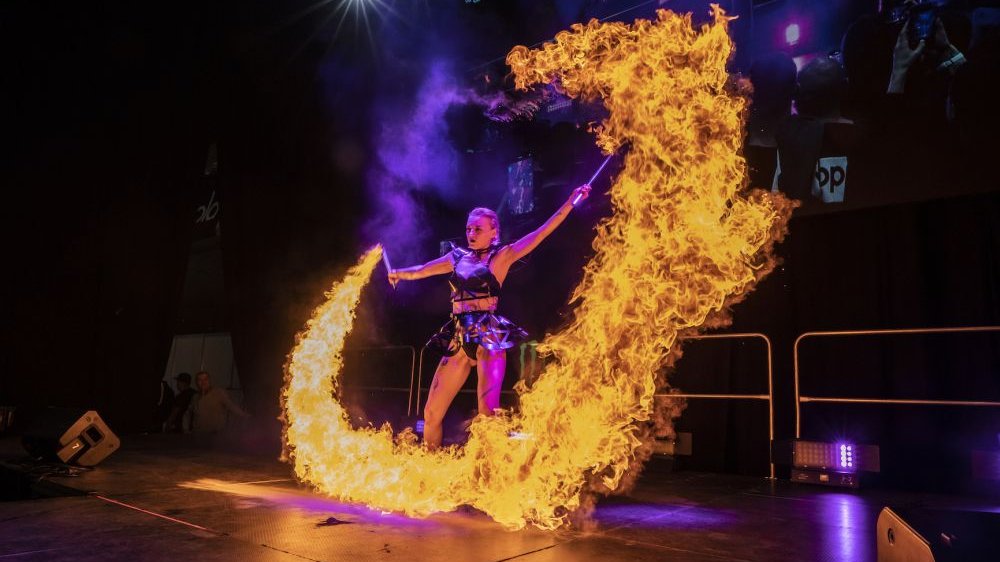 A woman in a leather outfit stands on the stage holding two torches in her hands, from which a huge fire bursts.
