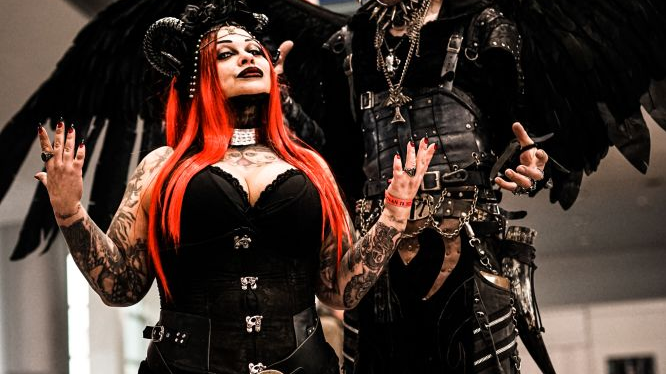 A couple in gothic costumes stand presenting their outfits. The woman has long, red hair and black horns on her head, the man has many earrings, big, black wings and diabolic makeup.