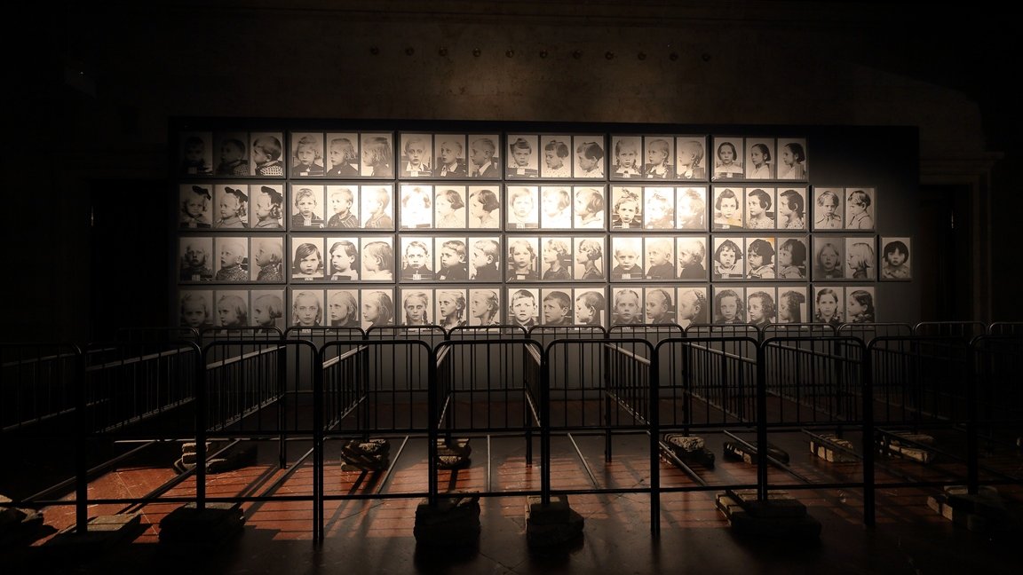 A photo from the exhibition: a dark room with old fashioned metal baby beds. In the background black and white portraits of children on a wall.