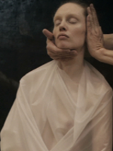 Photo of a women with her eyes closed, dressed in white cloth. Her face is being touched by someone's two hands.