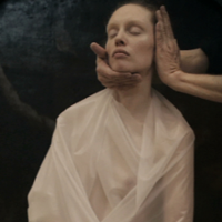 Photo of a women with her eyes closed, dressed in white cloth. Her face is being touched by someone's two hands.