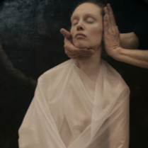 Photo of a women with her eyes closed, dressed in white cloth. Her face is being touched by someone's two hands.