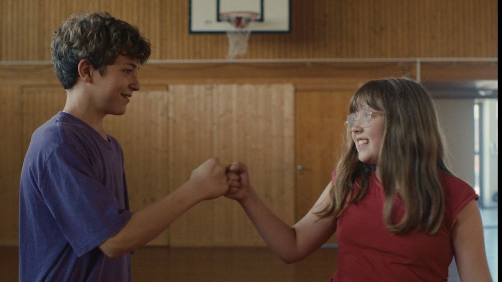 A boy and a girl who fistbump, in the background there is a wooden wall and a basketball hoop.