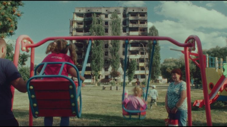 Photo of children swinging on two swings, two adilts near them. In a background destroyed block of flats.