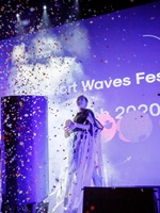 A woman standing on a stage. A big screen in the background. The picture in blue and violet colours, in front of the stage spread colourful confetti.