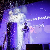 A woman standing on a stage. A big screen in the background. The picture in blue and violet colours, in front of the stage spread colourful confetti.