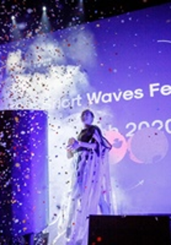 A woman standing on a stage. A big screen in the background. The picture in blue and violet colours, in front of the stage spread colourful confetti.
