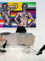 Photo of two men, one sitting at the desk, another opposite him in an arm-chair. Colourful picture on the wall as a background.