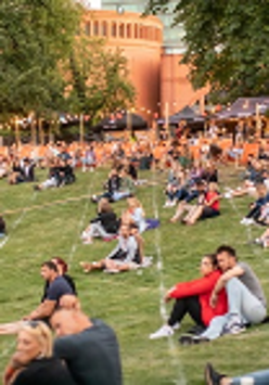 Colourful photo of people who sit on the grass in small groups, most often in pairs, and listen to a concert.