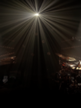 Photo of two artists performing on stage - one playing the piano, another playing drums. Black background and a single headlight lighting up the stage.
