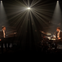 Photo of two artists performing on stage - one playing the piano, another playing drums. Black background and a single headlight lighting up the stage.