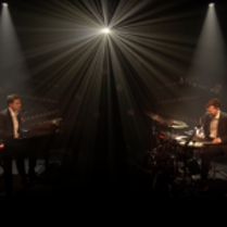 Photo of two artists performing on stage - one playing the piano, another playing drums. Black background and a single headlight lighting up the stage.