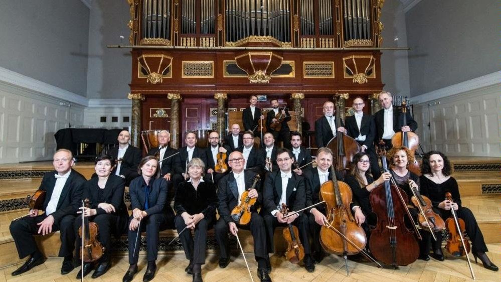 Photo of the orchestra - the musicians are sitting and standing in three rows on a stage, holding their instruments in their hands. The big pipe organ in the background