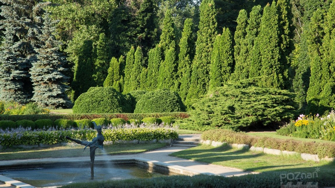Photo of a park with many green, trees and bushes of various hight lit by sunlight. In the foreground a fountain with a sculpture of a ballet dancer, surrounded with pavements.