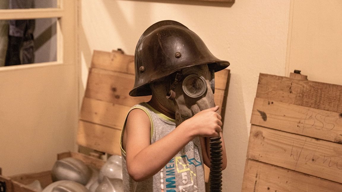 The picture presents a child in a gas-mask and a helmet. Wooden open chests in the background.