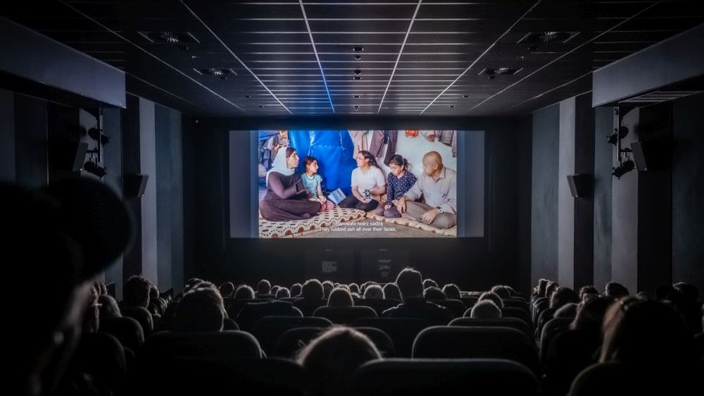 Photo of a cinema hall. In a foreground viewers sitting on arm-chairs in several rows, in a background - a cinema screen with a film displayed on it.