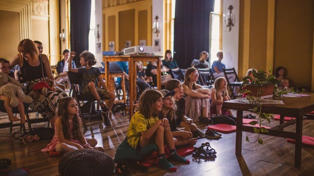 The room is full of children and parents, the children are sitting on poufs and pillows in the front row, looking carefully ahead.