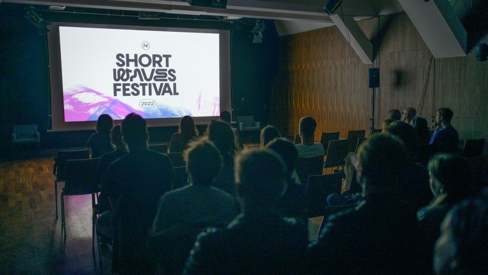 Dark room, a film show is going on, the Short Waves logo is visible on the screen. Spectators are sitting on chairs, looking at the screen.