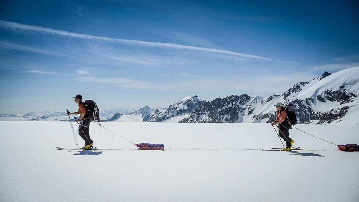 Photo from the movie - two persons skiing and pulling a baggage behind them. High mountains and blue sky as a background.