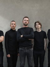 Photo of the band - five men in black clothes, grey wall as a background