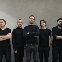 Photo of the band - five men in black clothes, grey wall as a background