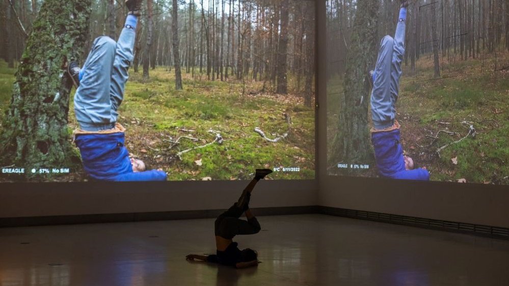 Photo from the performance - a dancer lying on the floor with her legs raised vertically. In the background, a fragment of a photo projected on the wall, showing a person in a similar pose on the grass by a tree.