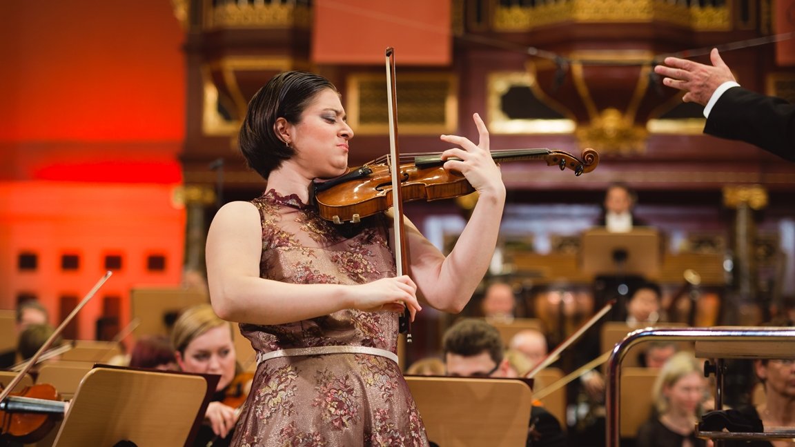 Photo of Veriko Tchumburidze playing the violin on stage. The musicians of the orchestra behind her.