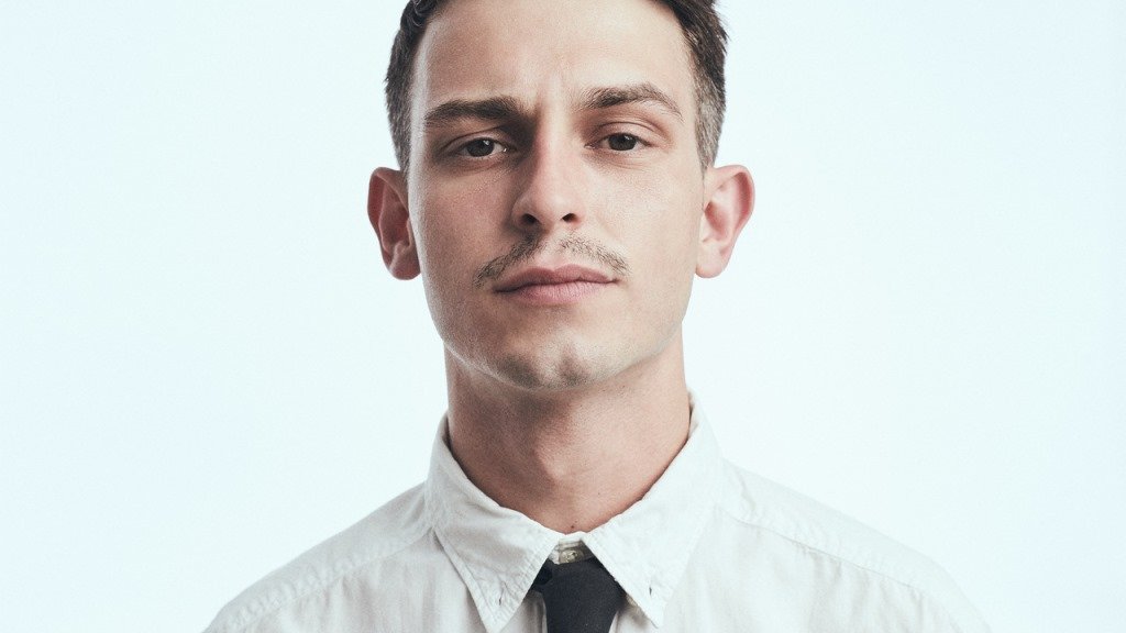 A picture of the artist - a young man in white shirt with black tie, who is looking straight into the camera