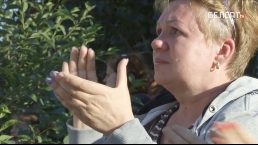 Picture from the movie - a woman who is clapping her hands and looking at something in front of her. Her face is serious. Some green bushes as a background.