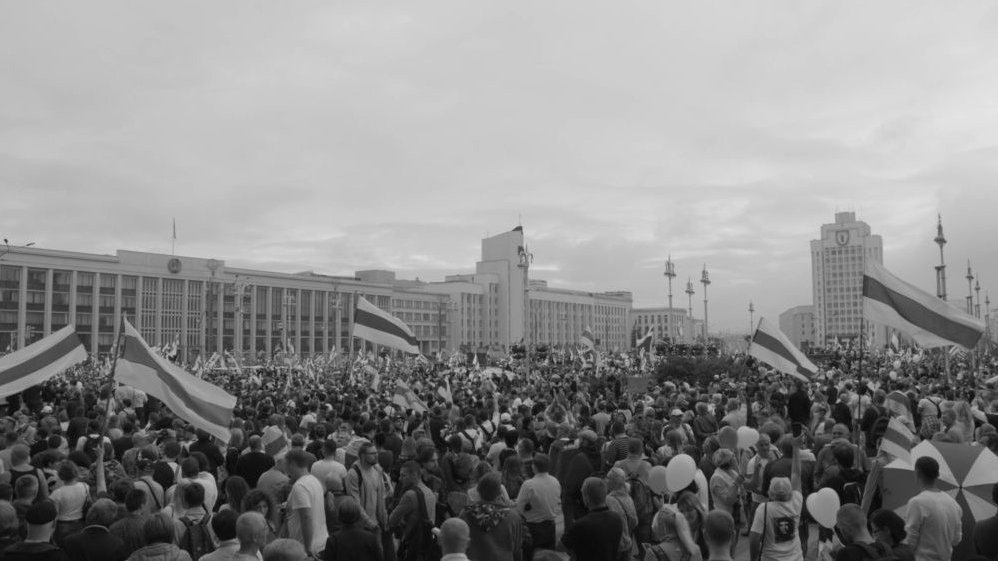 Black and white photo of a crowd of people in front of a white building which looks like an office building. Some people hold flags and balloons.