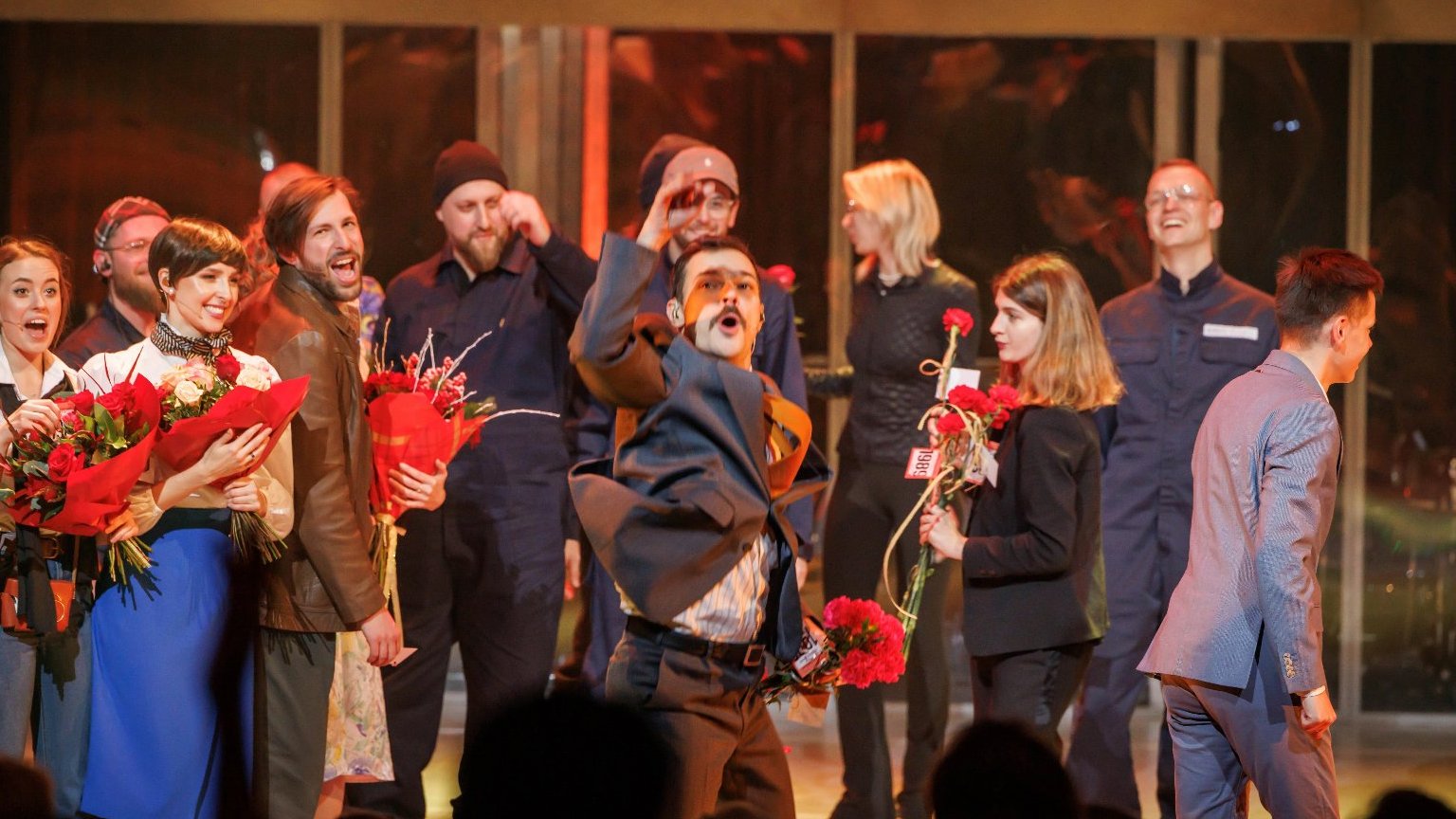 Kilka osób stoi na scenie, niketóre trzymają kwiaty. Mężczyzna z przodu unosi rękę w geście radości.