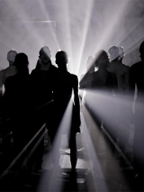 W ciemności stoi grupa taneczna, która jest oświetlona od tyłu bialym światłem.