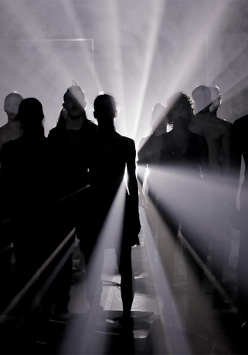 W ciemności stoi grupa taneczna, która jest oświetlona od tyłu bialym światłem.