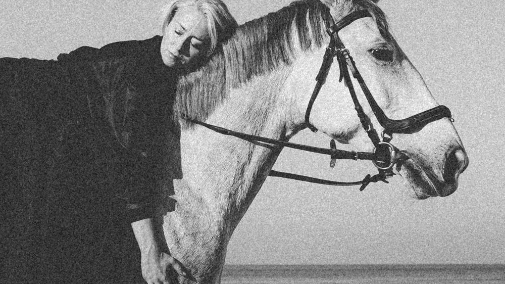 Artystka w czarnej szacie leży na białym koniu, przytulając się do jego szyi. Zdjęcie czarno-białe w staroświeckim klimacie.