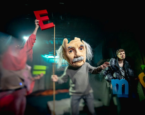 Aktor z przeskalowaną głową-maską Alberta Einsteina stoi na środku sceny.