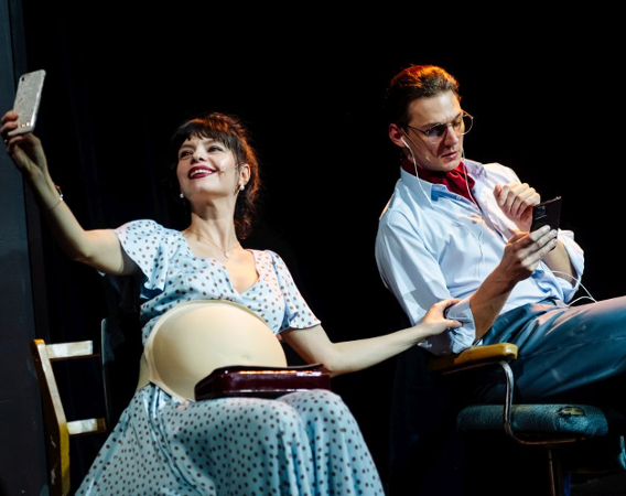 Aktorka ze sztucznym ciążowym brzuchem i aktor w okularach siedzą na krzesłach.