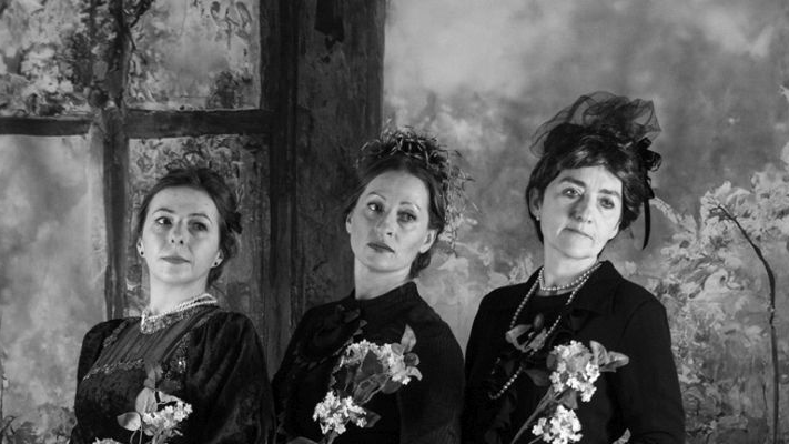 Czarno-biała fotografia stylizowana. Trzy kobiety w czarnych sukniach stoją w rzędzie, każda z nich trzyma w ręce kwiaty.