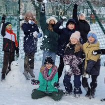 Grupa roześmianych dzieci pozuje do zdjęcia. Dzieci są ubrane w zimowe kurtki, czapki i rękawiczki. Bawią się śniegiem, podskakują i lepią bałwana.
