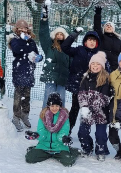 Grupa roześmianych dzieci pozuje do zdjęcia. Dzieci są ubrane w zimowe kurtki, czapki i rękawiczki. Bawią się śniegiem, podskakują i lepią bałwana.
