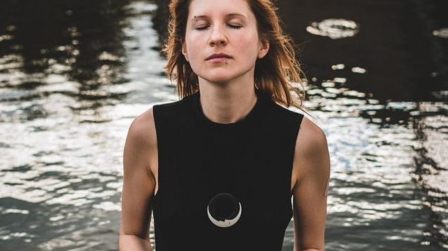 Kobieta w czarnej sukience stoi w wodzie trzymając w dłoniach instrument. Ma zamknięte oczy.