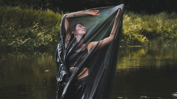 Kobieta w czarnym stroju stoi w wodzie owinięta półprzezroczystym płótnem.