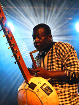 Mężczyzna w koszuli w kratę grający na korze - instrument przypomina gitarę. Za nim światła sceniczne.