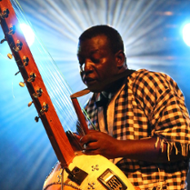 Mężczyzna w koszuli w kratę grający na korze - instrument przypomina gitarę. Za nim światła sceniczne.