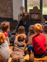 Zdjęcie, aktorka klęczy przed teatrzykiem postawionym na stole, przed nią siedzą i leżą dzieci.